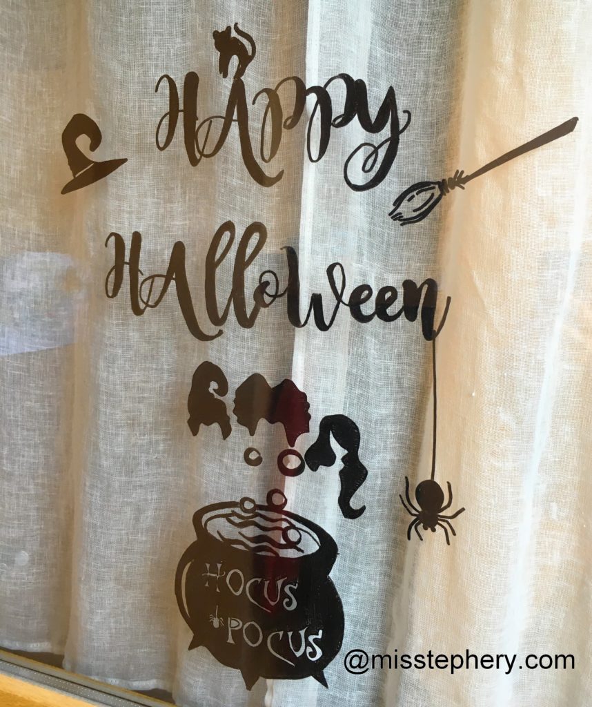 Dessin sur vitre au Posca noir d'un chaudron magique fumant et de l'inscription 'Hocus Pocus' pour Halloween, sur fond de rideau clair.
