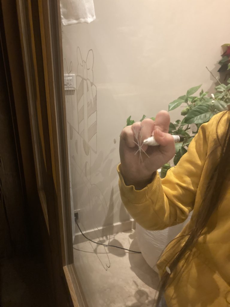 Tutoriel : comment dessiner sur une fenêtre avec un marqueur Posca pour Noël. La photo montre le traçage en cours d'un dessin de cadeau sur la vitre.