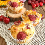 Trois muffins sans gluten et sans lactose aux cerises fraîches et amandes effilées, posés sur un napperon blanc à côté d'un bouquet de tournesols et de pivoines.