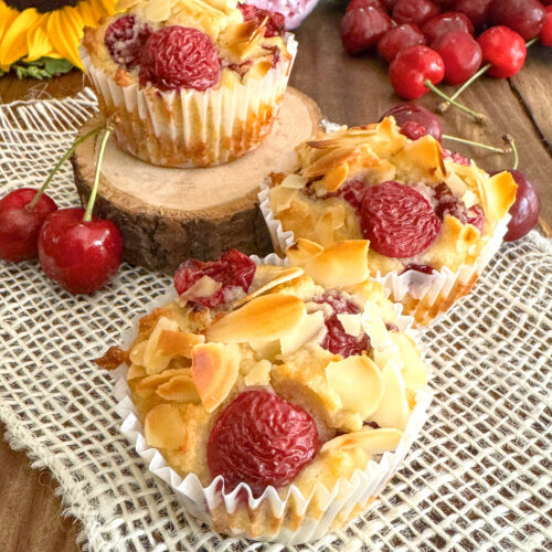 Trois muffins sans gluten et sans lactose aux cerises fraîches et amandes effilées, posés sur un napperon blanc à côté d'un bouquet de tournesols et de pivoines.