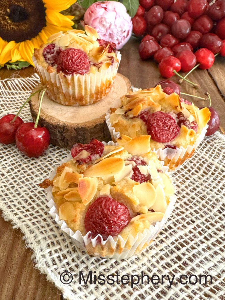 Trois muffins sans gluten et sans lactose aux cerises fraîches et amandes effilées, posés sur un napperon blanc à côté d'un bouquet de tournesols et de pivoines.