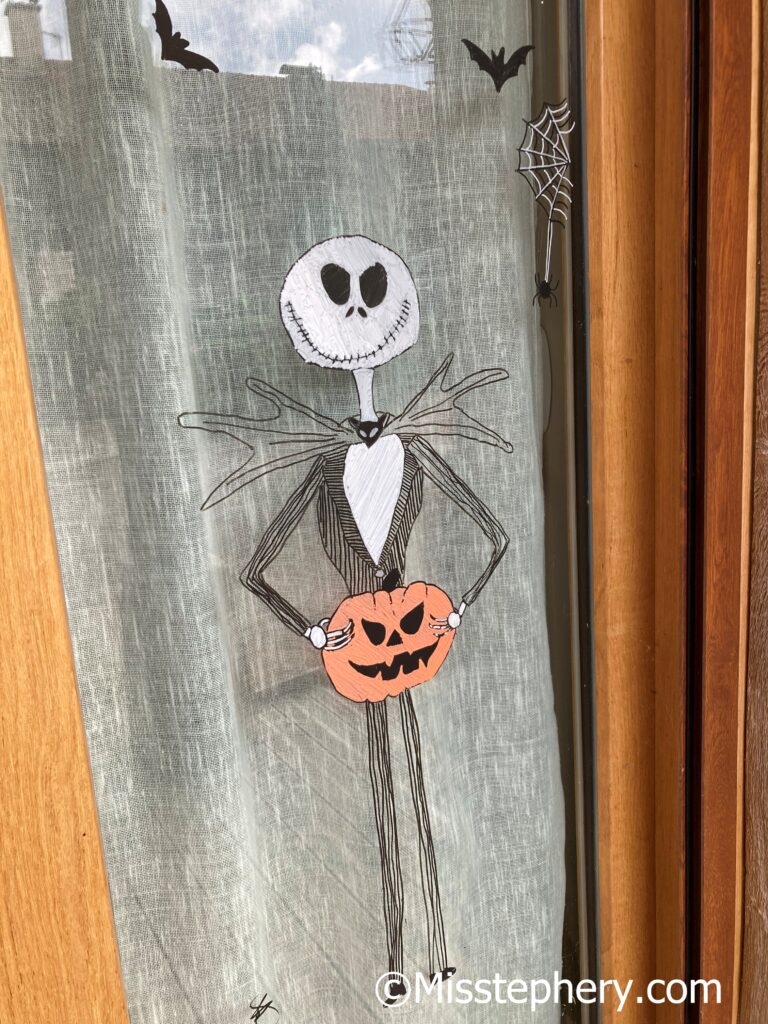 Dessin au Posca blanc et noir du personnage Jack Skellington tenant une citrouille d'Halloween orange sur une vitre.