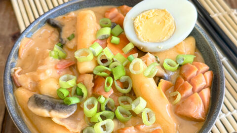 Rosé Tteokbokki : La version onctueuse, sans gluten ni lactose dans un bol composé de tteok, de saucisses de Francfort, de poitrine de fumés d'un demi oeuf dur posé sur le dessus parsemé de cébettes.