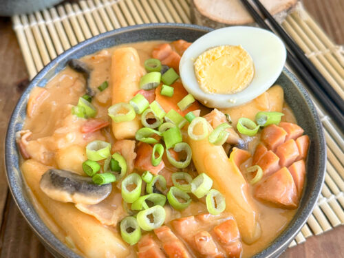 Rosé Tteokbokki : La version onctueuse, sans gluten ni lactose dans un bol composé de tteok, de saucisses de Francfort, de poitrine de fumés d'un demi oeuf dur posé sur le dessus parsemé de cébettes.