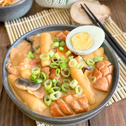 Rosé Tteokbokki : La version onctueuse, sans gluten ni lactose dans un bol composé de tteok, de saucisses de Francfort, de poitrine de fumés d'un demi oeuf dur posé sur le dessus parsemé de cébettes.