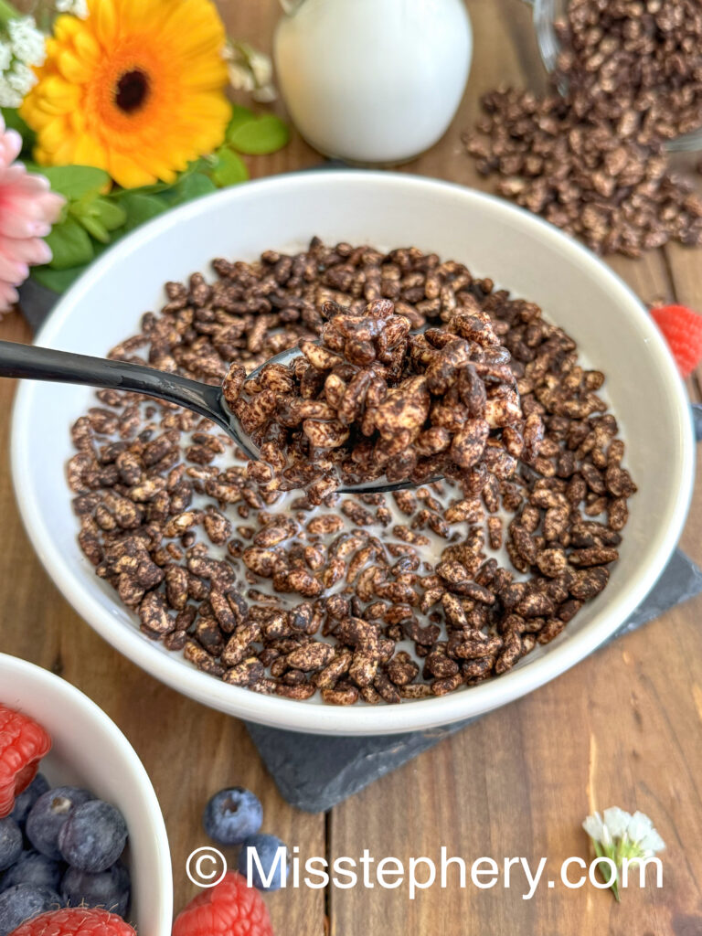 Une cuillère noire soulevant une bouchée croustillante de céréales au chocolat maison saines et de lait végétal au-dessus d'un bol.