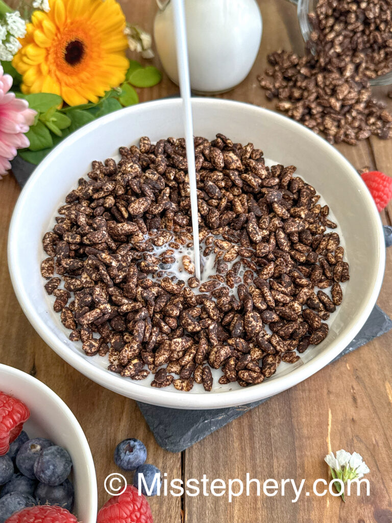 n filet de lait végétal versé dans un bol blanc rempli de céréales façon Coco Pops maison sans gluten et vegan, entouré de fruits rouges et de fleurs.