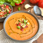 Un bol de gaspacho maison sans gluten rouge-orangé à la texture veloutée, décoré d'un filet d'huile d'olive, de dés de légumes croquants et de feuilles de basilic frais, sur un fond de tomates mûres et de poivrons.