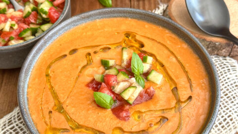 Un bol de gaspacho maison sans gluten rouge-orangé à la texture veloutée, décoré d'un filet d'huile d'olive, de dés de légumes croquants et de feuilles de basilic frais, sur un fond de tomates mûres et de poivrons.