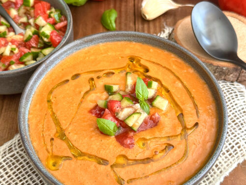 Un bol de gaspacho maison sans gluten rouge-orangé à la texture veloutée, décoré d'un filet d'huile d'olive, de dés de légumes croquants et de feuilles de basilic frais, sur un fond de tomates mûres et de poivrons.