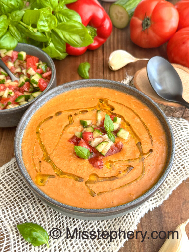 Un bol de gaspacho maison sans gluten rouge-orangé à la texture veloutée, décoré d'un filet d'huile d'olive, de dés de légumes croquants et de feuilles de basilic frais, sur un fond de tomates mûres et de poivrons.
