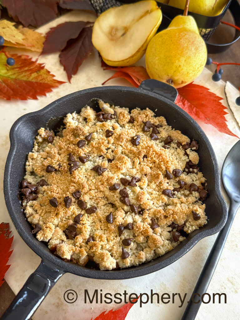 Crumble d'automne 100% végétal aux poires et chocolat noir sans gluten, dans une poêle en fonte, entouré de décoration automnale.