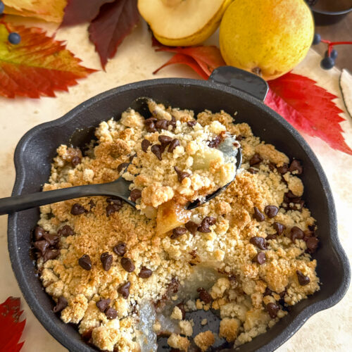 Une bouchée de crumble végétalien aux poires et pépites de chocolat, dessert d'automne servi chaud dans une mini poêle en fonte sur un fond décoratif de feuilles et de fruits.