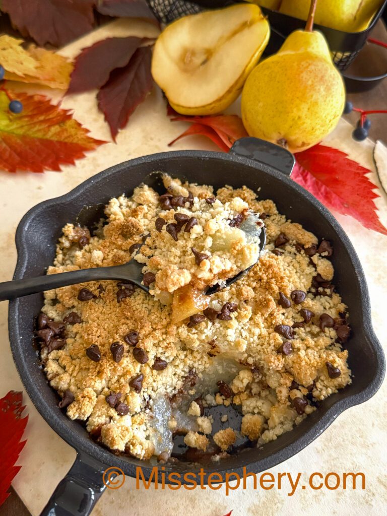 Une bouchée de crumble végétalien aux poires et pépites de chocolat, dessert d'automne servi chaud dans une mini poêle en fonte sur un fond décoratif de feuilles et de fruits.